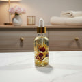 A hand holds a small glass dropper bottle filled with golden oil and dried red flower petals, with a blurred kitchen background.